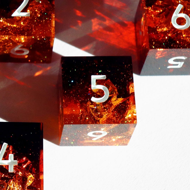 May include: Close-up of several dice with a fiery, glittery interior. The dice are transparent with white numbers, including the number 5. The overall color scheme is orange and red, with a bright white background.