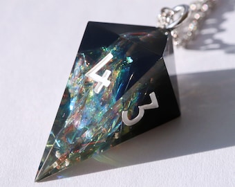 Könnte beinhalten: Eine schwarze, geometrische Anhänger-Halskette mit einer silbernen Kette. Der Anhänger hat eine dunkle, transparente Form mit schillernden, farbenfrohen Einschlüssen. Silberne Zahlen "4" und "3" sind auf der Oberfläche des Anhängers sichtbar. Die Halskette wird auf weißem Hintergrund präsentiert.