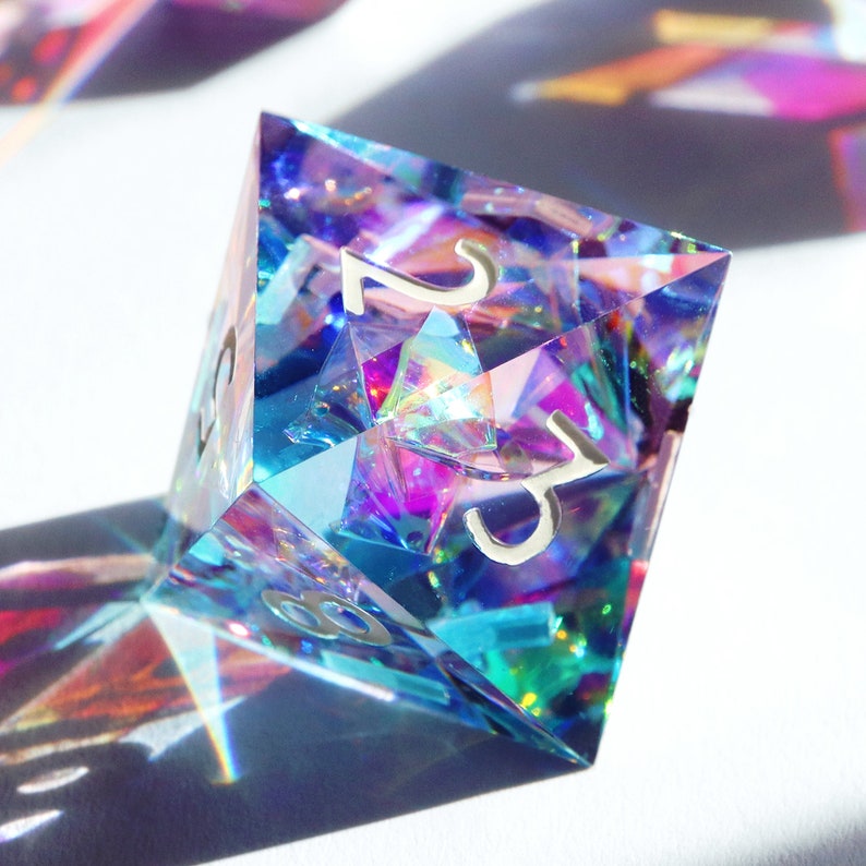 May include: A iridescent blue and purple crystal d20 dice with silver numbers. The dice is a polyhedral die with 20 sides.
