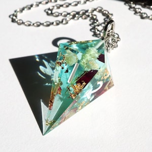 Könnte beinhalten: Ein tealblauer Harz-D20-Würfel-Anhänger mit einer silbernen Kette. Der Würfel hat getrocknete Blumen und Blattgold eingebettet. Die Zahl 2 ist auf einer der Seiten sichtbar.