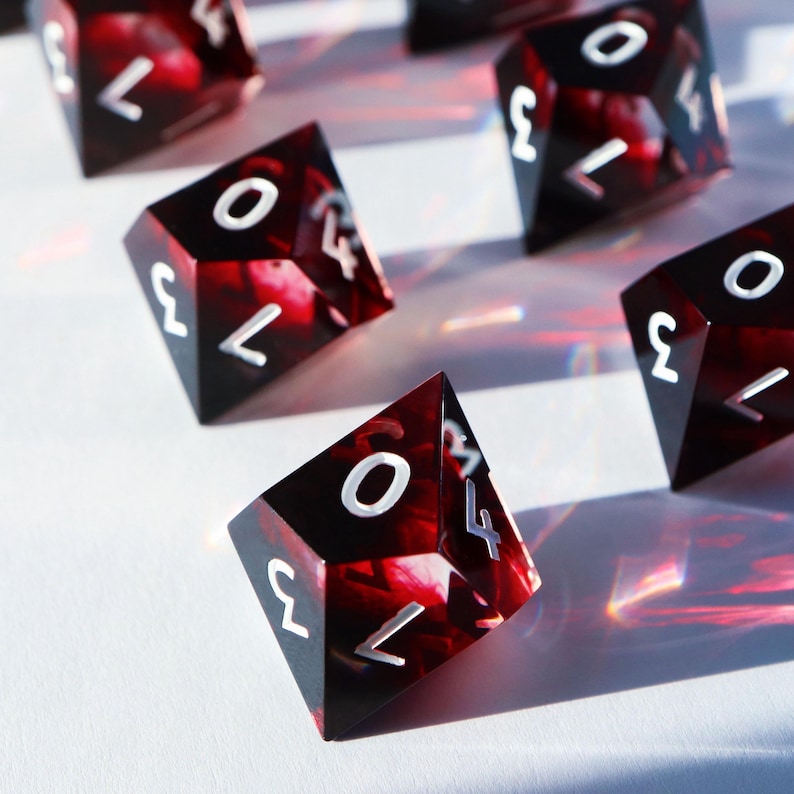 May include: Several polyhedral dice with a marbled red and black design. The dice have white numbers and symbols. The dice are scattered on a white surface.