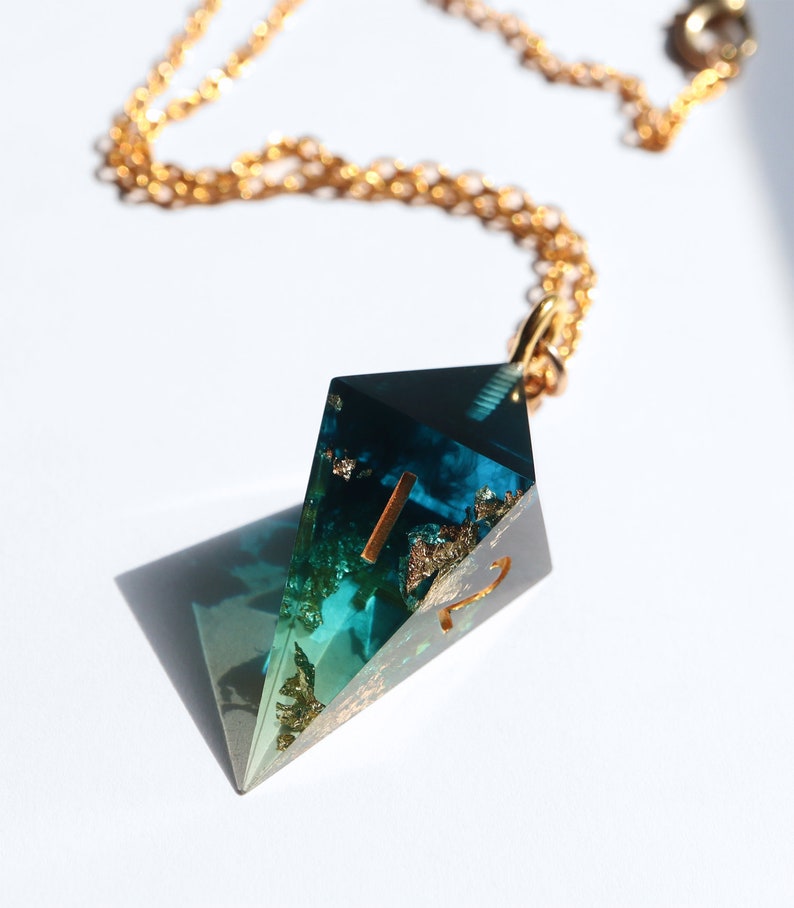 Puede incluir: Collar con colgante geom&eacute;trico y cadena dorada. El colgante es de resina transparente con inclusiones de hojas de oro y tonos turquesa. Forma de diamante.