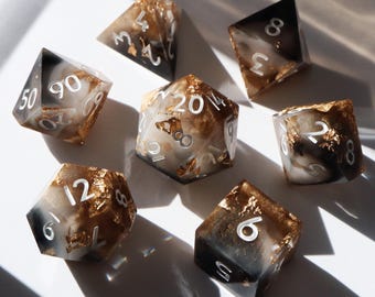 May include: Set of seven polyhedral dice with black and white marbled resin and gold leaf accents. The dice are numbered 1-20, 4, 6, 8, 12, and 50.