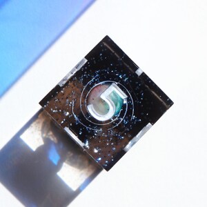 May include: A square, black resin object featuring the number "5" within a circle. The number is white with green and red detailing. The resin is speckled with blue glitter. The object casts a shadow on a white surface.