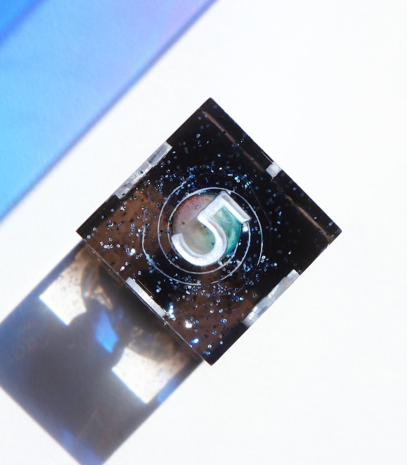 May include: A square, black resin object featuring the number "5" within a circle. The number is white with green and red detailing. The resin is speckled with blue glitter. The object casts a shadow on a white surface.
