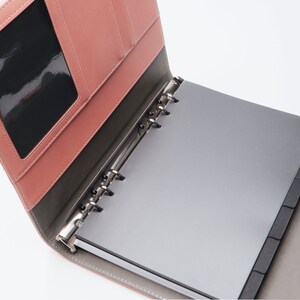 May include: A pink leather bound notebook with a clear plastic window on the front cover. The notebook has a silver ring binder and black dividers.