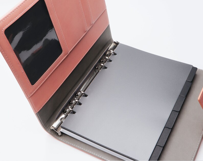 May include: A pink leather bound notebook with a clear plastic window on the front cover. The notebook has a silver ring binder and black dividers.