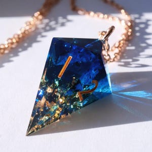 Puede incluir: Un collar con colgante geométrico con un diseño de resina azul. El colgante tiene detalles dorados y los números 1 y 2. La cadena del collar es dorada.