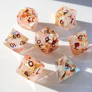 May include: Set of seven clear resin dice with gold foil and iridescent flakes. Each die has black numbers. The dice are in various geometric shapes, including a d20, d12, d10, d8, d6, and d4. The dice are for tabletop games.