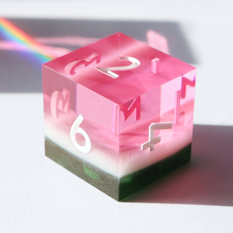 May include: A pink, white, and green layered acrylic dice with white numbers. The dice is showing the number 6.