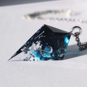 May include: A geometric pendant necklace with a silver chain. The pendant is a clear, angular shape with a dark blue and silver interior. The letter "R" is visible within the pendant.