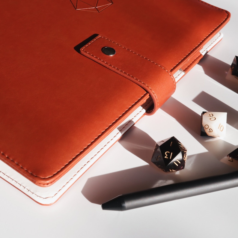 May include: A brown leather journal with a white stitched edge and a geometric design on the cover. The journal is open to reveal a blank page. There are several dice and a black pen on a white surface next to the journal.