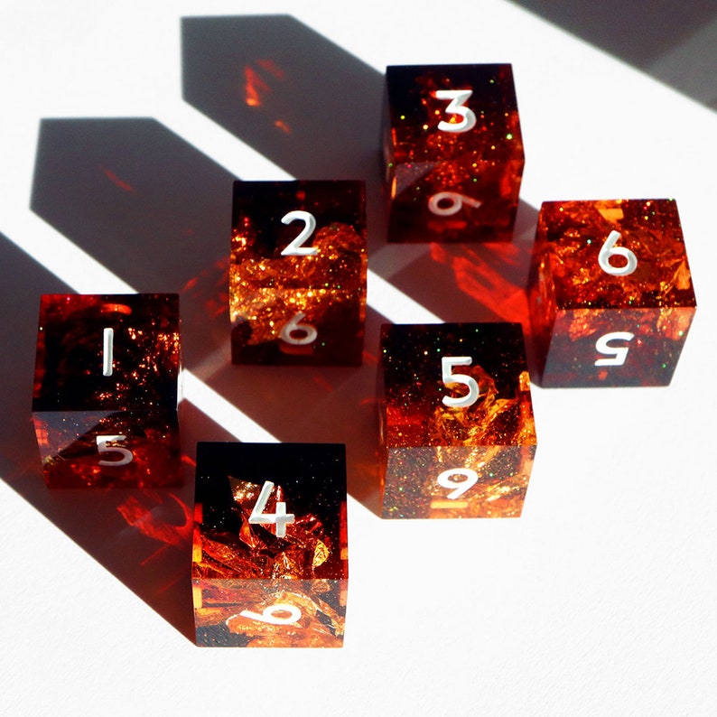 May include: Six square dice with a fiery, amber-colored interior and white numbers. The dice are arranged on a white surface, with shadows cast across the scene. The dice are numbered 1 through 6.