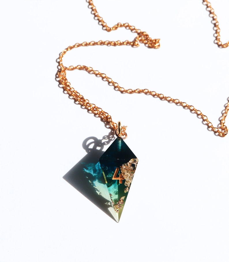 Puede incluir: Un collar de cadena dorada con un colgante geom&eacute;trico. El colgante tiene forma de diamante con un dise&ntilde;o turquesa y dorado, y el n&uacute;mero 4 en dorado. El collar est&aacute; sobre un fondo blanco.