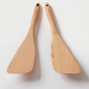 May include: Two wooden spatulas with light brown tones. Each spatula has a wide, angled head and a long handle with a small hole at the top. The spatulas are made of wood and are designed for cooking.