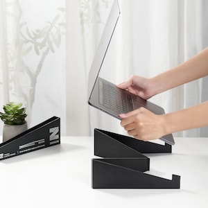 Puede incluir: Un soporte para portátil negro con un portátil encima. El soporte está hecho de dos piezas que están conectadas entre sí. El portátil está abierto y la pantalla está mirando hacia la cámara. El soporte está sobre una mesa blanca.