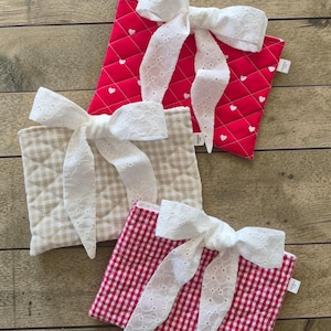 Peut inclure: Trois pochettes en tissu matelassé avec des nœuds en dentelle blanche. L'une est rouge avec des cœurs blancs, une autre est beige et blanche à carreaux, et la troisième est rouge et blanche à carreaux. Les pochettes sont disposées sur une surface en bois.