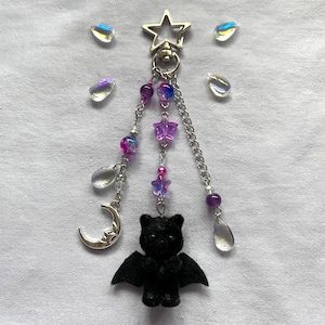 May include: A silver keychain featuring a star clasp, a black bat charm, purple and pink beads, a crescent moon charm, and iridescent teardrop accents. This gothic-style keychain is a unique accessory.