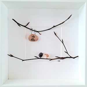 Peut inclure: Une œuvre d'art encadrée représentant deux petits oiseaux peints perchés sur une branche avec une pierre brune suspendue à une autre branche au-dessus d'eux. La pierre porte les mots "Elle & Roby" écrits dessus.
