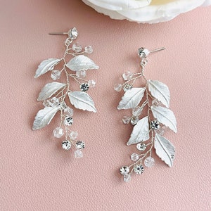 May include: Silver leaf earrings with clear crystal accents. The earrings are dangling and feature a delicate, leafy design.