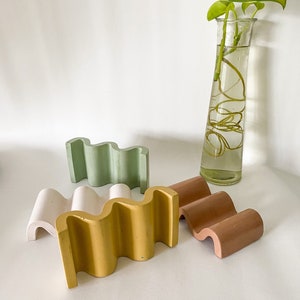 Puede incluir: Una colección de bandejas de hormigón con forma de onda en varios colores: rosa pálido, amarillo, verde y marrón. Un jarrón de cristal transparente con una planta verde en el fondo. Las bandejas están diseñadas para guardar objetos pequeños.
