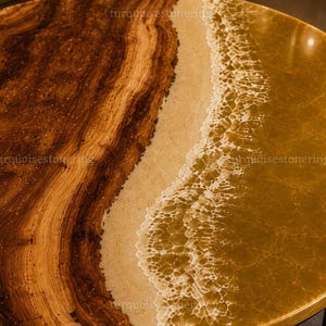 Puede incluir: Una mesa redonda con una losa de madera natural con vetas prominentes en un lado, que transiciona hacia una franja de arena texturizada y una secci&oacute;n de resina dorada transl&uacute;cida que imita olas con detalles de espuma blanca.