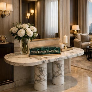 Puede incluir: Una consola con tapa de mármol y dos patas cilíndricas. Un gran espejo con marco dorado cuelga sobre la mesa. Un jarrón con flores blancas y una caja decorativa con el texto "TURQUOISESTONERING" están sobre la mesa. La habitación tiene una estética lujosa y elegante.