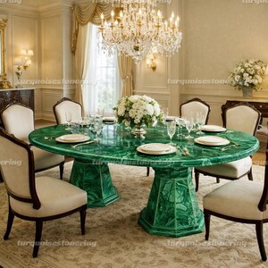 Può includere: Un tavolo da pranzo ovale in malachite verde smeraldo con base a piedistallo abbinata. Il tavolo è apparecchiato con piatti bianchi, posate e bicchieri. Sedie color crema con finiture in legno scuro circondano il tavolo. Un grande lampadario è appeso sopra.