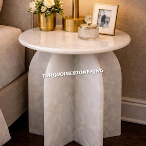 May include: A round marble side table with a unique base design. The table features a white marble top and a base with a cross-like structure. A gold lamp, a small gold vase with flowers, and a decorative jar are on the table. Text: TURQUOISESTONERING.