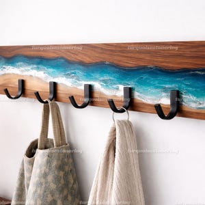 Può includere: Un attaccapanni da parete in legno con cinque ganci in metallo nero. Il legno è tinto di un marrone caldo e presenta un design in resina che raffigura una scena di spiaggia con onde blu e bianche. Due borse e una sciarpa sono appese ai ganci.