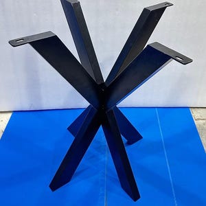 Puede incluir: Una base de mesa de metal negro que presenta un eje central con cuatro patas anguladas que se extienden hacia afuera. Cada pata tiene una placa de montaje plana en la parte superior con una ranura preperforada para fijar el tablero.