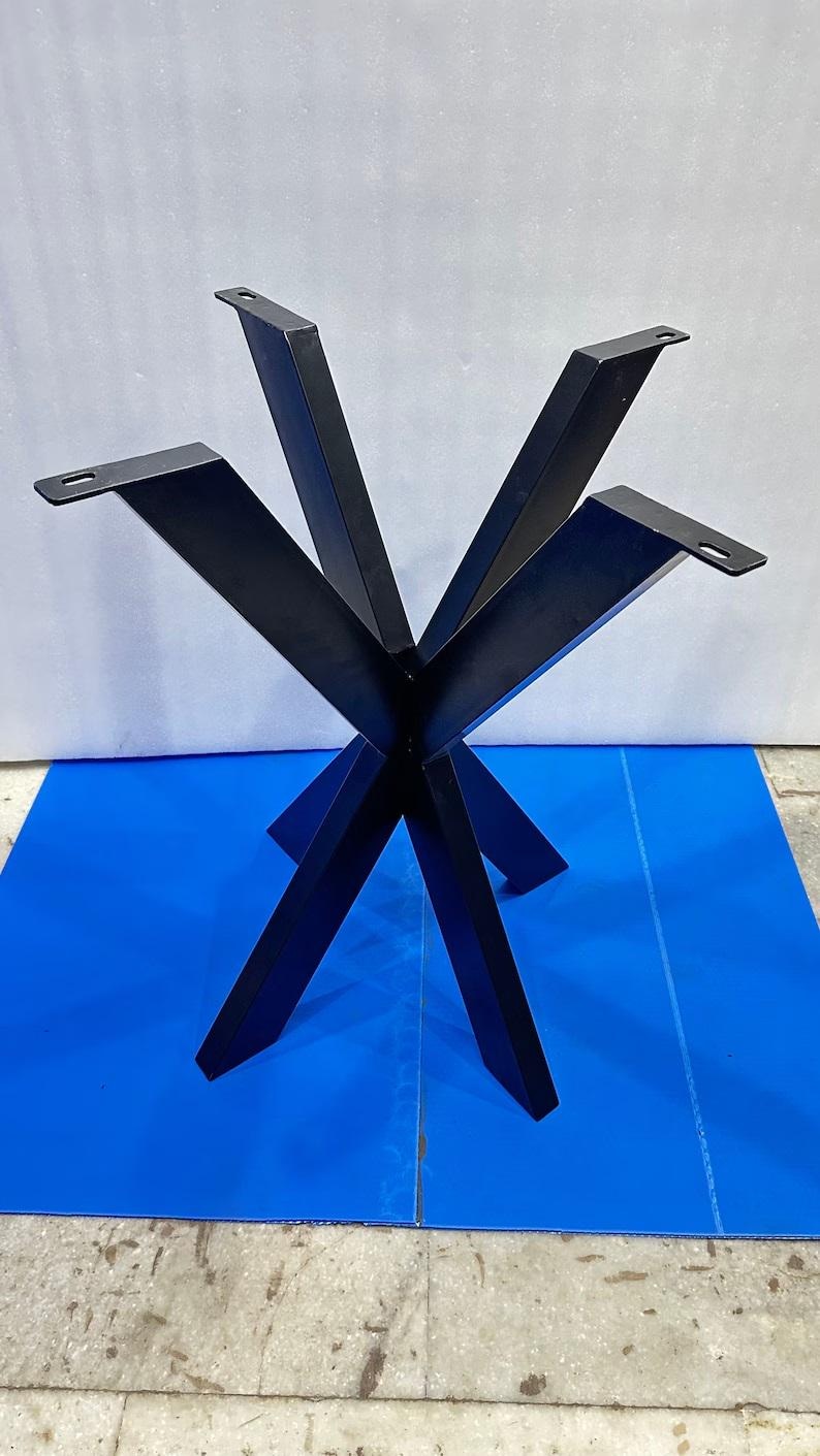 Puede incluir: Una base de mesa de metal negro que presenta un eje central con cuatro patas anguladas que se extienden hacia afuera. Cada pata tiene una placa de montaje plana en la parte superior con una ranura preperforada para fijar el tablero.