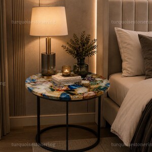 May include: A round side table featuring a multicolored agate stone tabletop with blue, white, and amber tones, supported by a thin black metal frame. The table holds a lamp with a cylindrical base, a small candle, and a decorative plant.