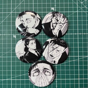 Puede incluir: Cinco botones redondos en blanco y negro de estilo manga, con diferentes ilustraciones de una persona en varios estados emocionales. Los botones tienen un acabado brillante. El fondo es una esterilla de corte verde.