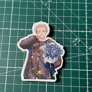 May include: A sticker featuring a cartoon character holding a bouquet of blue flowers. The character is wearing a brown jacket with star-shaped accents. The sticker is on a green cutting mat with grid lines.