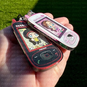 May include: Two acrylic keychains shaped like vintage mobile phones. One is black and red with the text JESSE and PINKMAN, featuring a cartoon character. The other is white and pink with the text SAULGOODMAN, featuring a cartoon character.