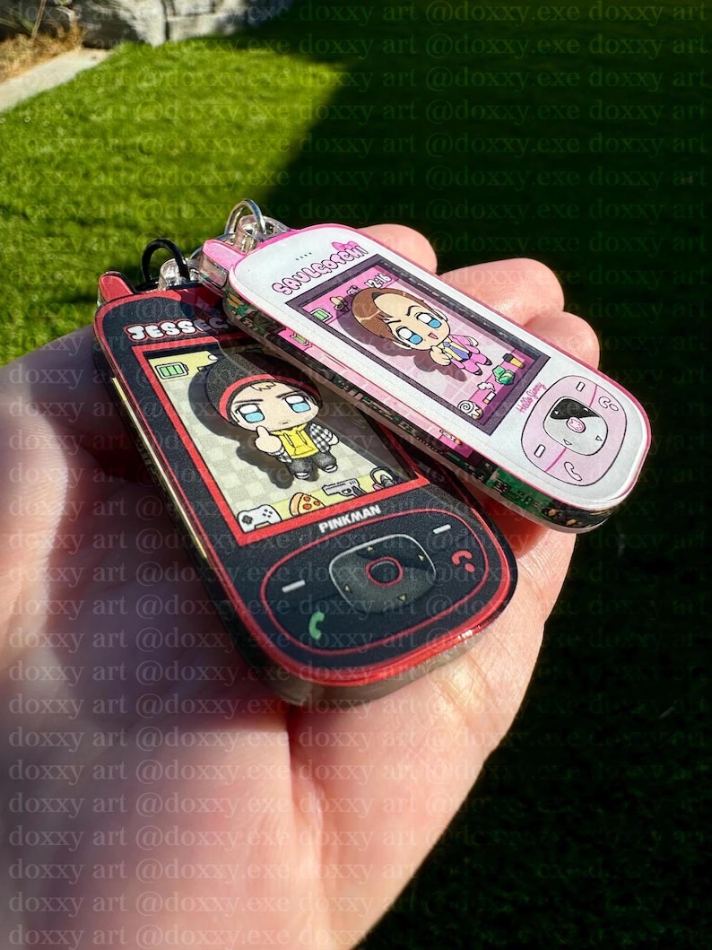 May include: Two acrylic keychains shaped like vintage mobile phones. One is black and red with the text JESSE and PINKMAN, featuring a cartoon character. The other is white and pink with the text SAULGOODMAN, featuring a cartoon character.