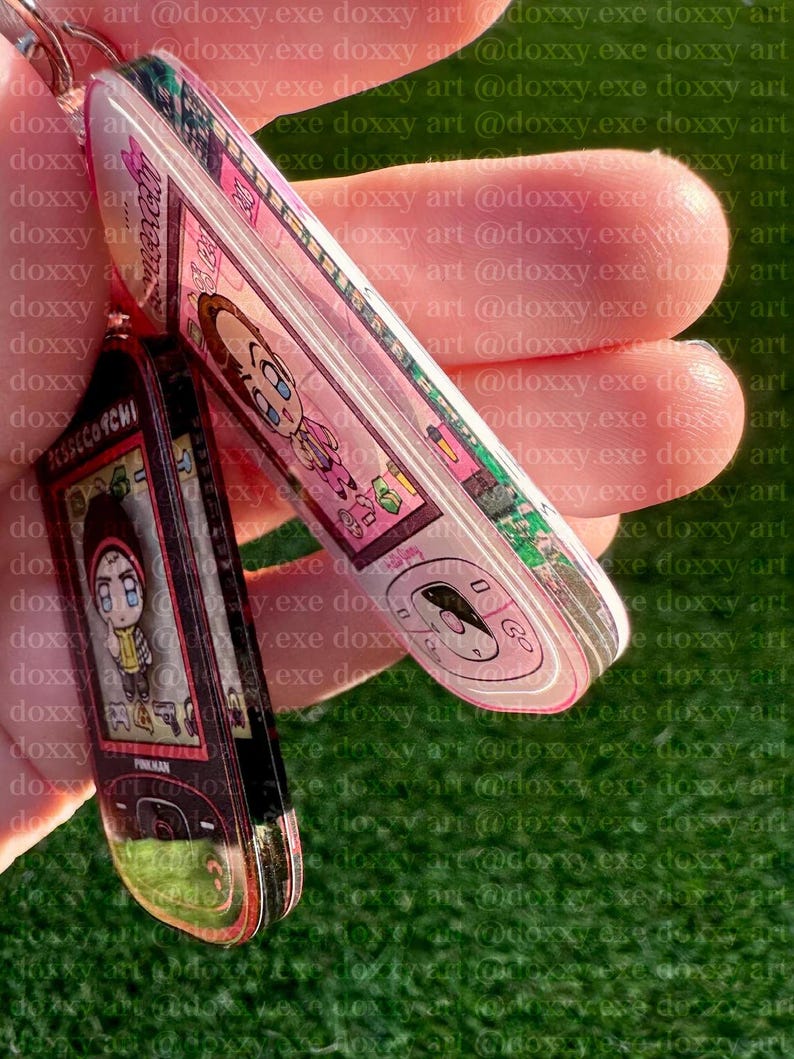 May include: Two acrylic keychains shaped like retro mobile phones. One is pink with a cartoon character and the text 'Jesse', the other is black with a cartoon character and the text 'Pinkman'. Both feature digital screen graphics and buttons.
