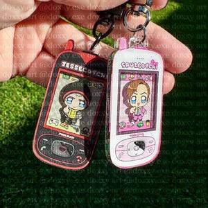 May include: Two acrylic keychains shaped like vintage mobile phones. The left keychain is black and red with the text JESSECOTCHI and PINKMAN, featuring a cartoon character. The right keychain is white and pink with SAULGOTCHI and Hello Jimmy.