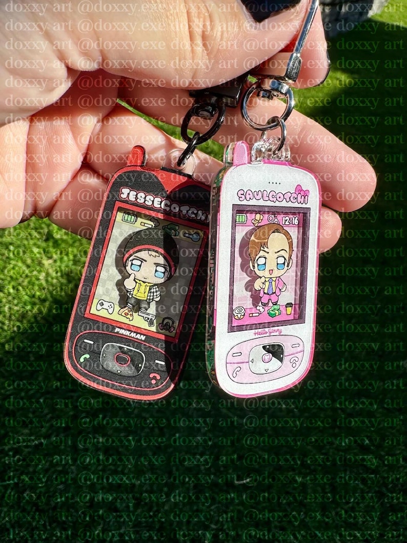 May include: Two acrylic keychains shaped like vintage mobile phones. The left keychain is black and red with the text JESSECOTCHI and PINKMAN, featuring a cartoon character. The right keychain is white and pink with SAULGOTCHI and Hello Jimmy.