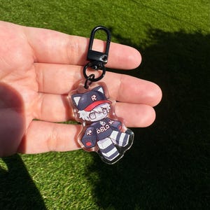 May include: A black and white cartoon character keychain with a red baseball cap and the word "ROBLOX" on the shirt. The keychain is attached to a black metal ring.