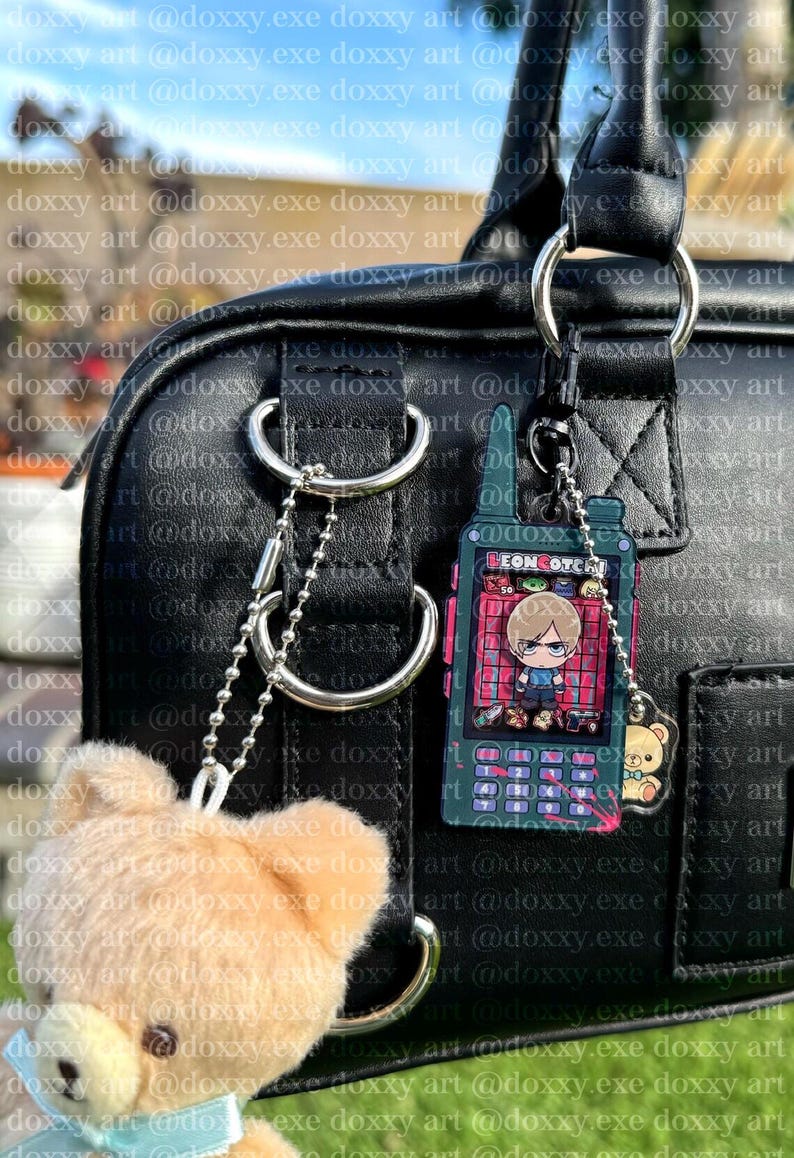 K&ouml;nnte beinhalten: Eine schwarze Tasche aus Kunstleder mit einem silbernen Metallring. Am Ring befestigt ist ein dunkelgr&uuml;ner, rechteckiger Anh&auml;nger in Form eines Funkger&auml;ts mit dem Text LEONCOTCH und einer Comicfigur sowie ein kleiner brauner Teddyb&auml;r-Anh&auml;nger.