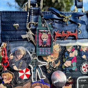 K&ouml;nnte beinhalten: Eine dunkelblaue Ita-Tasche, verziert mit verschiedenen Anh&auml;ngern, Anstecknadeln und Schl&uuml;sselanh&auml;ngern zum Thema Resident Evil. Zu den Details geh&ouml;ren ein zentraler radiof&ouml;rmiger Anh&auml;nger, ein roter You Are Dead-Pin und ein Umbrella-Logo.