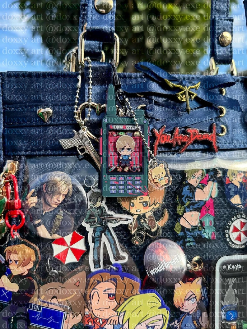 K&ouml;nnte beinhalten: Eine dunkelblaue Ita-Tasche, verziert mit verschiedenen Anh&auml;ngern, Anstecknadeln und Schl&uuml;sselanh&auml;ngern zum Thema Resident Evil. Zu den Details geh&ouml;ren ein zentraler radiof&ouml;rmiger Anh&auml;nger, ein roter You Are Dead-Pin und ein Umbrella-Logo.