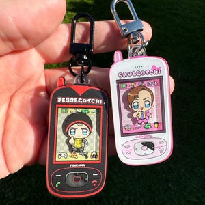 Puede incluir: Dos llaveros de teléfono celular rosa y negro con personajes de dibujos animados. Los llaveros presentan el texto "Jessecotch" y "Saulgotchi" con las palabras "Pinkman" y "Hello Skinny" en la parte inferior de los llaveros.