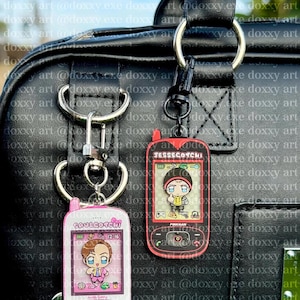 May include: Two rectangular acrylic keychains shaped like vintage mobile phones. The left keychain is pink with the text SAULGOTCHI and a cartoon character. The right keychain is red with the text JESSEGOTCHI and a cartoon character with the name PINKMAN.