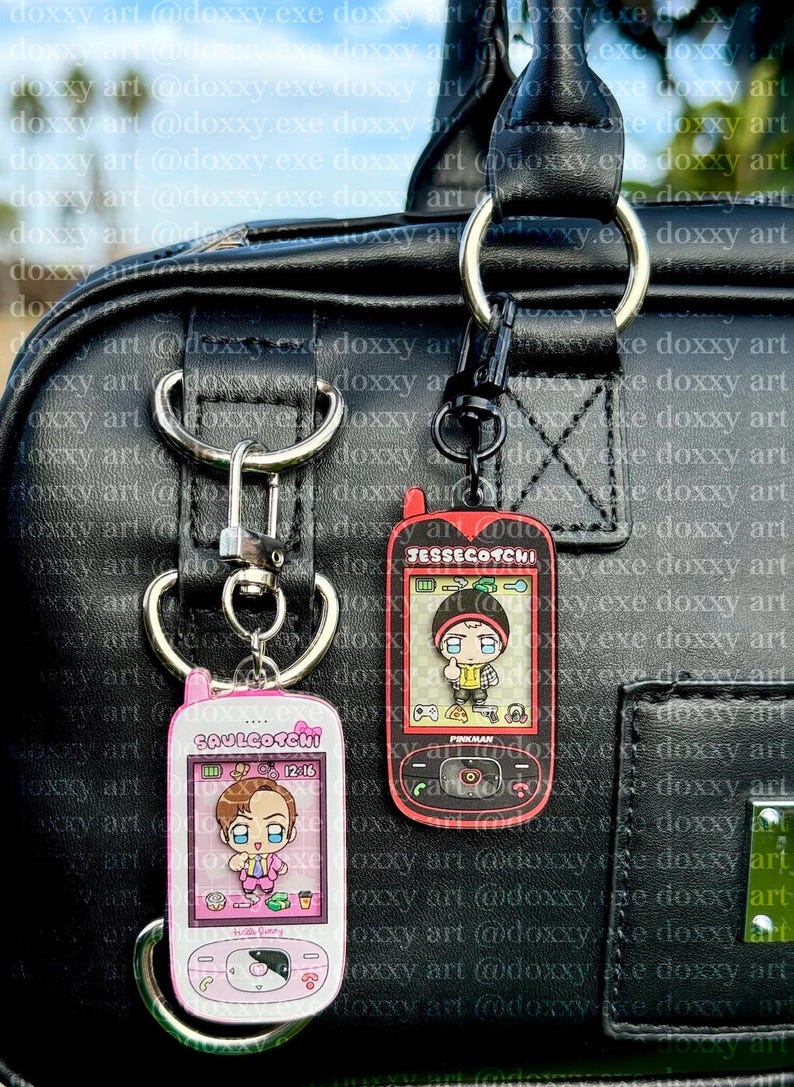 May include: Two rectangular acrylic keychains shaped like vintage mobile phones. The left keychain is pink with the text SAULGOTCHI and a cartoon character. The right keychain is red with the text JESSEGOTCHI and a cartoon character with the name PINKMAN.