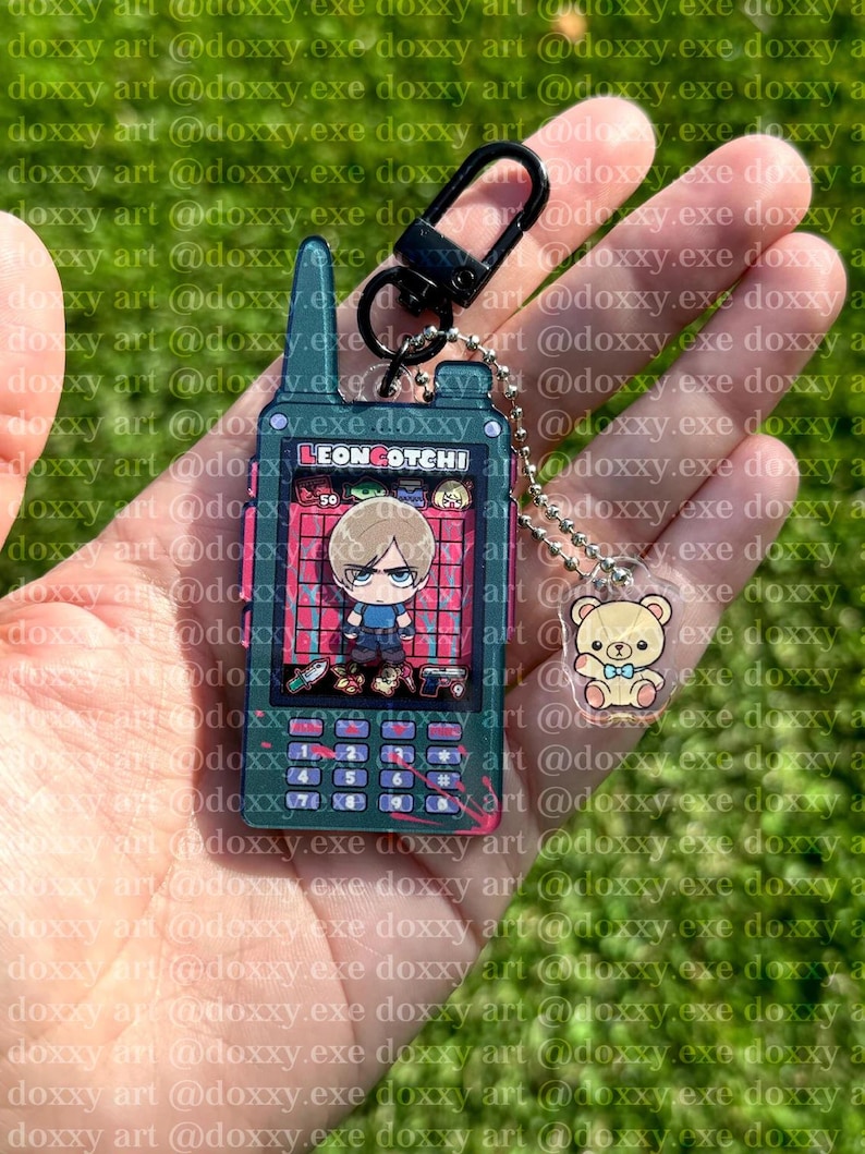 K&ouml;nnte beinhalten: Ein dunkelgr&uuml;ner Schl&uuml;sselanh&auml;nger in Form einer tragbaren Spielkonsole mit dem Text LEONGOTCHI auf dem Bildschirm, einer Zeichentrickfigur mit blonden Haaren und einem kleinen B&auml;renanh&auml;nger an einer silbernen Kugelkette mit schwarzem Karabiner.