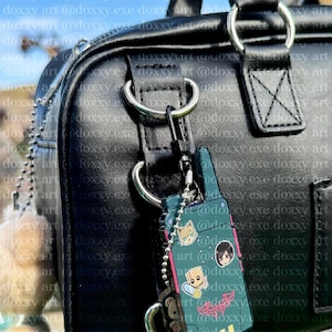 K&ouml;nnte beinhalten: Eine schwarze Tasche aus Kunstleder mit einem dunkelgr&uuml;nen, rechteckigen, funkger&auml;tf&ouml;rmigen Schl&uuml;sselanh&auml;nger. Der Anh&auml;nger zeigt drei kleine Cartoon-Aufkleber und ein rosa Grafikdesign und ist mit einer silbernen Kugelkette befestigt.
