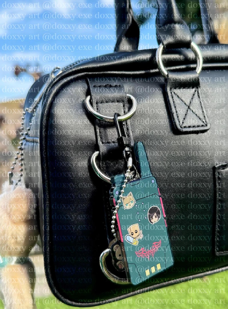 K&ouml;nnte beinhalten: Eine schwarze Tasche aus Kunstleder mit einem dunkelgr&uuml;nen, rechteckigen, funkger&auml;tf&ouml;rmigen Schl&uuml;sselanh&auml;nger. Der Anh&auml;nger zeigt drei kleine Cartoon-Aufkleber und ein rosa Grafikdesign und ist mit einer silbernen Kugelkette befestigt.